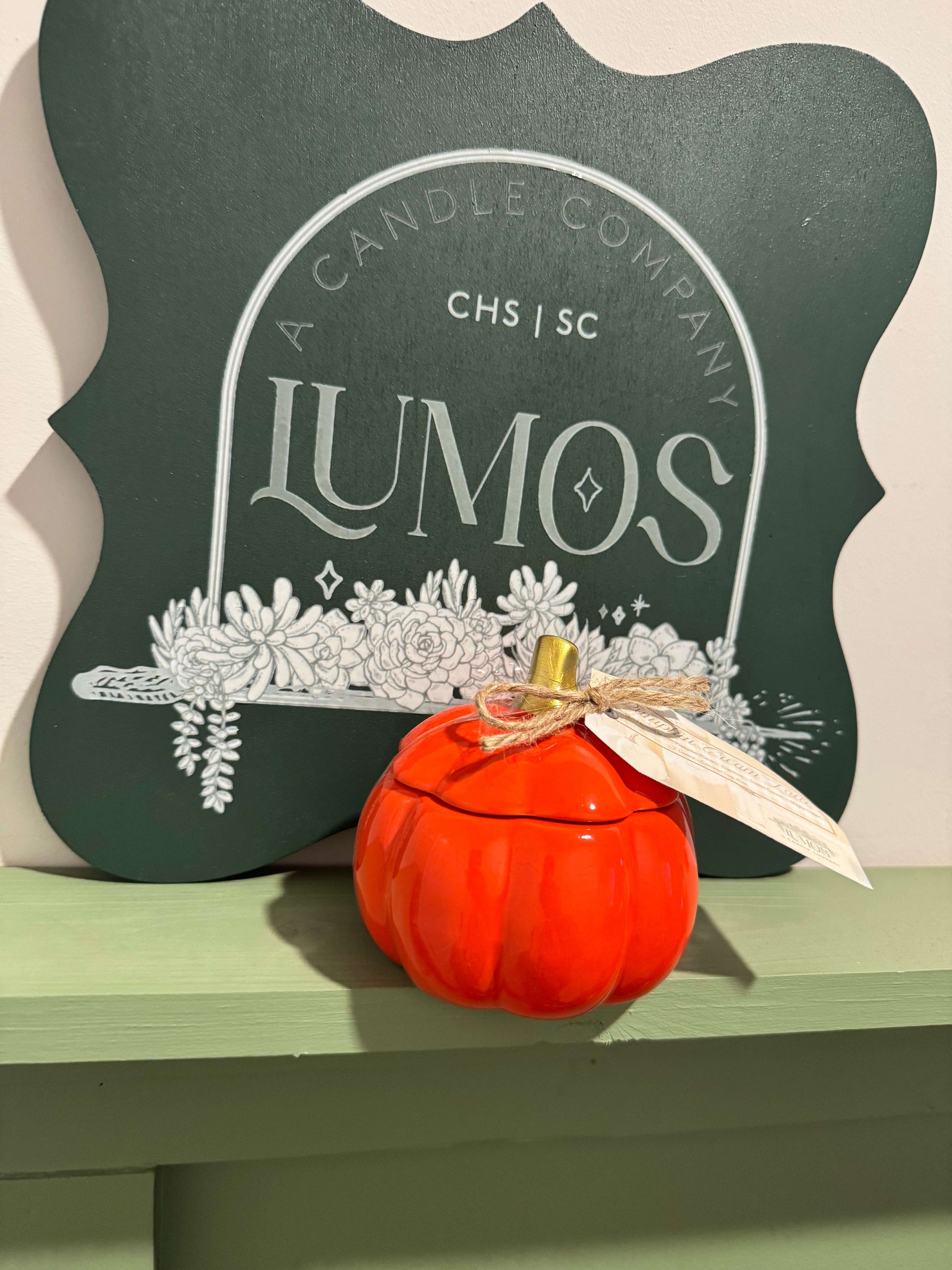 Fall scented candle in orange pumpkin ceramic vessel with bow and tag displayed on green shelf in front of Lumos logo sign