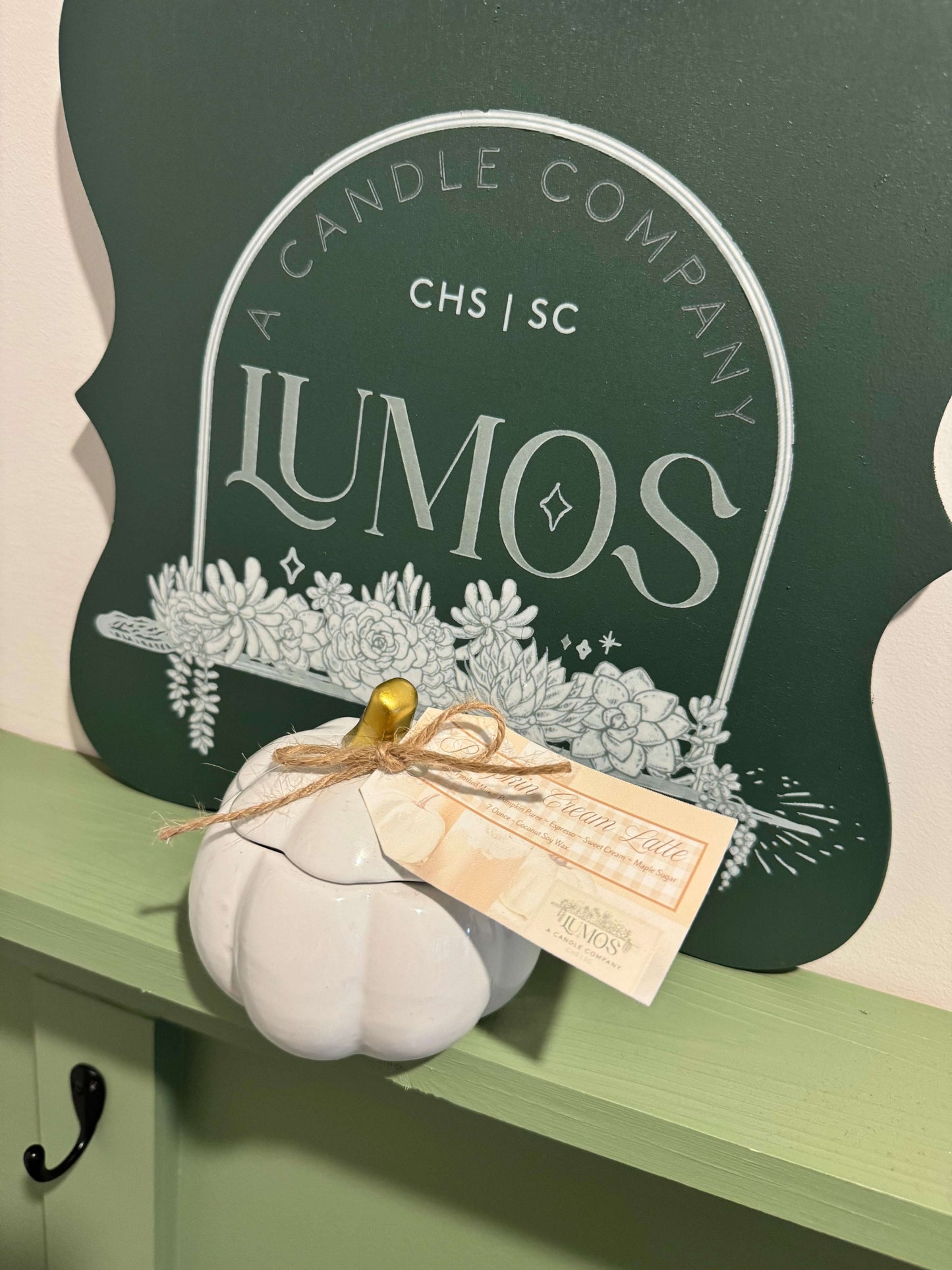 Fall scented candle in white pumpkin ceramic vessel with gold stem and twine detail displayed on green shelf with Lumos sign background