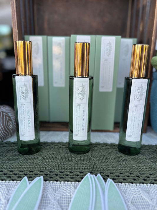 LUMOS Room + Linen Spray luxury home fragrance mist in elegant glass bottles with gold sprayers on a green and white textured surface
